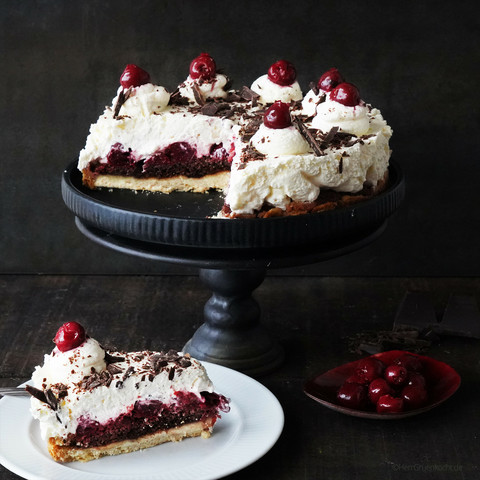 
Dunkler Hintergrund. Auf einer Kuchenetagere eine Schwarzwälder-Kirsch-Tarte. Im Vordergrund ein Stück davon auf einem weißen Teller.