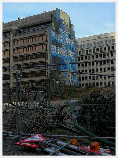 Ein Baustellenzaun in Brüssel vor einer Brachfläche und dem Gebäude mit dem großen Wandgemälde "The Future is Europe". Vor dem Bauzaun liegen abgebaute Verkehrsschilder mit Ampel-Symbol, 
vom Bildvordergrund verläuft über den Bauschutt eine grüne Leitung Richtung europäische Zukunft-Wandbild. 