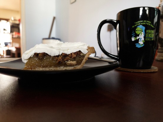 A slice of pecan pie with whip cream on top sits atop a square purple plate. The plate sits on a desk, next to it is a mug of coffee sitting on a coaster. There is an alien in pajamas, holding a cup of coffee, on the coffee mug. Words on the mug say “Mornings are rough… No matter where you’re from.”