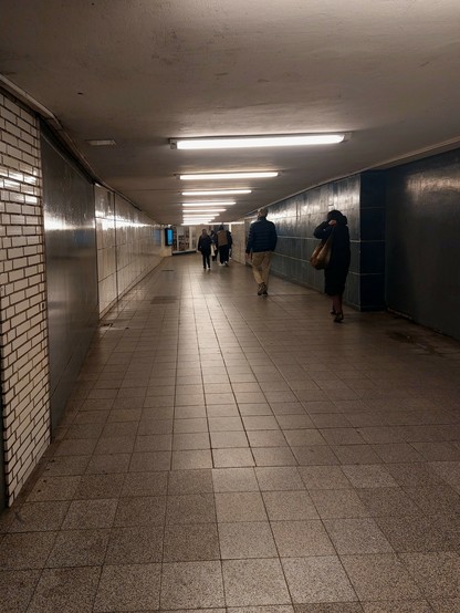 Ein trostloser mit Steinplatten gepflasterter Gang mit gefliesten Wänden, links weiß, rechts dunkelblau, später auch weiß. Darüber eine Reihe quer angebrachter Neonröhren. Mehrere Personen hasten durch den Gang Richtung Ausgang.