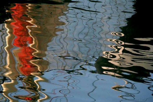 Liggend beeld. Links: van bovenaf taps aflopende crème kleurige vorm, beweeglijk, met fel rode baan in het midden.
Midden: spiegeling van de blauwe lucht met grillige verticale lijntjes.
Rechts: driehoekige zwarte vorm, onderin het breedst. Met ook onderin sierlijke crème kleurige ronde oogvormige spiegelingen. 
