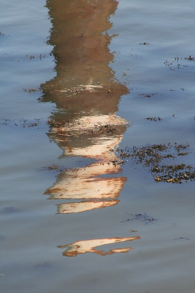 Spiegeling van een bruine roestige meerpaal, met witte top, in blauw water. 
Op het water drijft een beetje blaaswier.