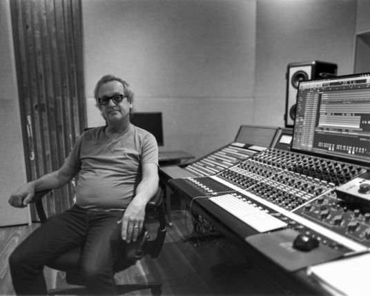 A man with a bemused expression sits next to a vintage Neve console in a recording studio control room. He is wearing dark jeans and a light t shirt and dark rimmed glasses. He is being patient with the photographer. 