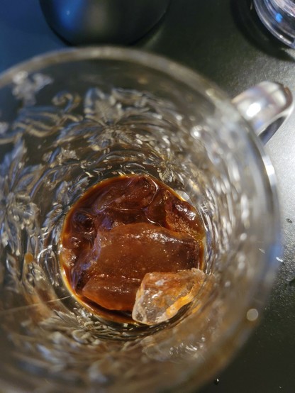 Looking down at a deep rich chocolate brown liquid with ice cubes in a decorative clear mug, sitting on a black desk.