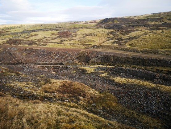 A view across a disused lead mine landscape. In the bottom of the valley is the river Nent, which is walled on both sides and the area both sides of the river is rocky mine waste. Moorland rises above and the sky is grey. 
