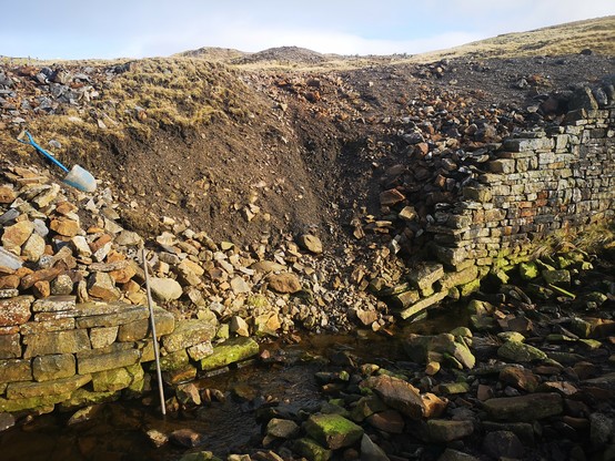 An area of collapsed wall and riverbank. Stones from the wall are in the river and lead mine waste has fallen into the river, leaving a large, fan shaped depression in the bank. Length of the repair is about 4 metres. 