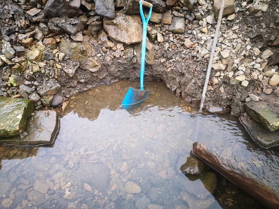 After digging out the base of a collapsed riverbank wall, a pool has formed. In it are a blue spade and a steel pinch bar. Water is about 6 inches deep. 