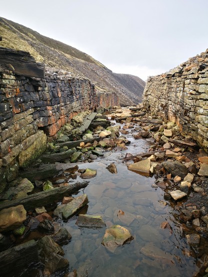 A view looking downstream between the walls lining the river Nent. The walls are up to 2 metres high. There are several serious bulges and gaps in the stonework. Some rotting timbers are visible from the time when the river bed was fully lined in wood. 