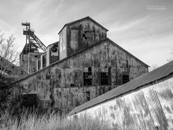 Black and white view of the rusty metal crusher building at an abandoned lead mine with the head house seen in the background.