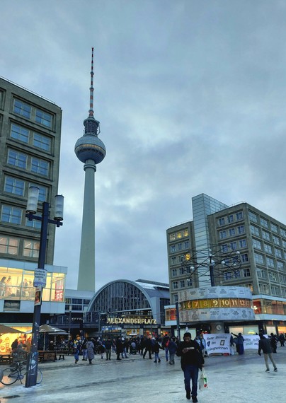 Foto über den östlichen Teil des Alexanderplatz mit Blick auf den S-Bahnhof, dahinter der Fernsehturm und davor die Weltzeituhr. Es sind nicht übermäßig viele Menschen unterwegs, der Himmel ist grau bewölkt. An der Weltzeituhr stehen Menschen mit dem Banner "Keine Stimme den Klimaleugnerinnen" Eine Person trägt eine Papperde in Herzform mit der Aufschrift "Omas for Future".