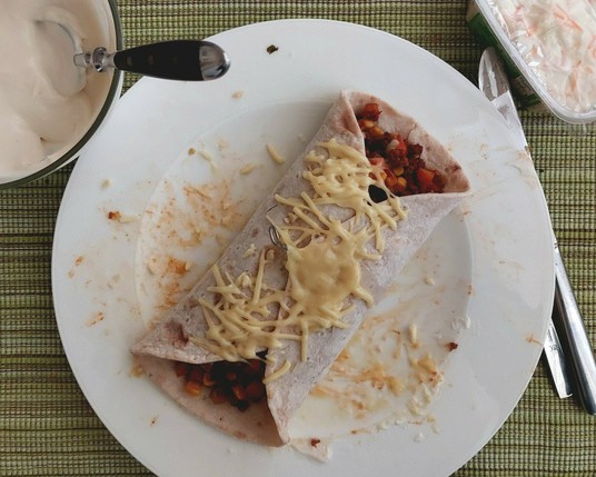 Ein Tortilla mit der beschriebenen veganen Füllung und mit Käse überbacken auf rinem weißen Teller. Daneben eine Schüssel Joghurt und ein Becher veganer Cole Slaw.