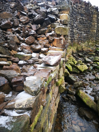 Looking along a dry stone wall that rises from the bed of a river. Further away is original height, about 2.5m. Nearer, the wall is being rebuilt and will be a little lower. Lead Mine waste infills behind the wall. There is a dusting of snow on some of the stones. 