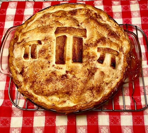 Color image of an apple pie with the Greek symbol for Pi baked into the crust. The image shows the apple pie with three Pi symbols, a browned domed crust, on a metal cooling rack over a red gingham tablecloth.