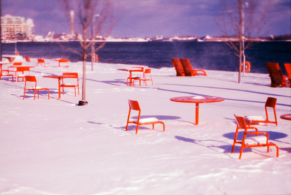 Bright red metal furniture in frozen in crisp bright snow as the wind whips the snow in a haze off the edge of the pier. 