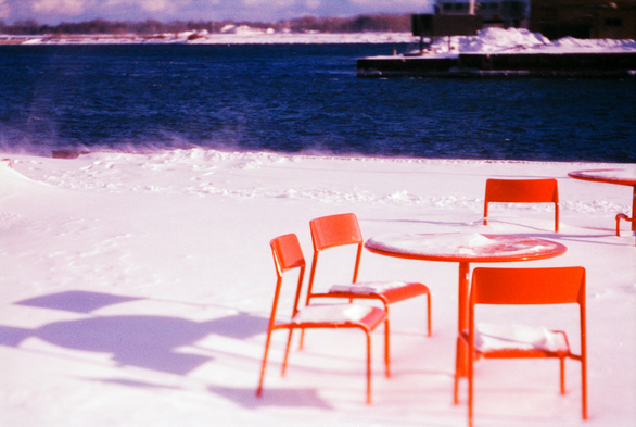 The bases of a red table and chairs are locked in the snow. Closer to the edge of the pier, where the wind forms tails of snow over the lake, another chair is buried over its seat. 