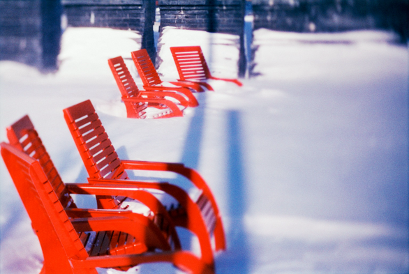 Two groups of three red metal Muskoka chairs are locked in the snow as the dark bulk of the Irish Famine Memorial rises in sections beyond. 