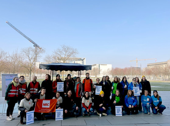 Eine Gruppe von Menschen, darunter Mitglieder von Terre des Hommes, posiert gemeinsam für ein Foto. Sie halten Schilder mit Fakten zur Krisenprävention und Armut in den Händen. Einige tragen farbige Westen oder Mützen. Im Hintergrund ist das Bundeskanzleramt in Berlin zu sehen, sowie Baukräne und Bäume. Der Himmel ist klar und blau.