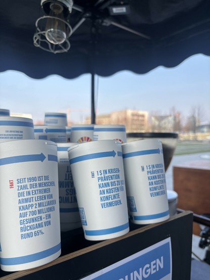 Mehrere weiße Pappbecher mit blauen Aufdrucken stehen gestapelt auf einem Tresen. Auf den Bechern stehen Fakten zur weltweiten Armutsbekämpfung und Krisenprävention in deutscher Sprache. Ein Fakt besagt, dass die Zahl der Menschen in extremer Armut seit 1990 von fast 2 Milliarden auf 700 Millionen gesunken ist. Im Hintergrund sind verschwommene Personen und ein blauer Himmel sichtbar.