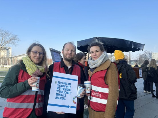Drei Personen tragen rote Westen mit dem Logo von Terre des Hommes. Eine Person hält ein Schild mit der Aufschrift: „Seit 2000 hat sich die weltweite Kindersterblichkeit mehr als halbiert.“ Im Hintergrund sind weitere Menschen und ein Stand zu sehen.