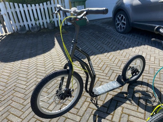 An urban scooter with a black frame, two wheels, and a textured footboard is positioned on a patterned pavement. A car is partially visible in the background, along with a white picket fence. Bright sunlight casts shadows on the ground.