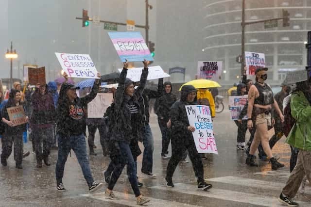 A group of protesters march in heavy rain, holding various signs with messages about gender rights and safety. Some participants wear rain gear, while others appear in summer clothing. Umbrellas are present, and the atmosphere looks energetic despite the downpour.