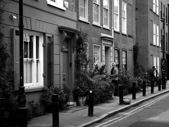 Black and white. A man is standing outside a block of flats on the left side of a road in East London. He has just knocked on the door and/or rung the bell and is standing back, waiting. Large planters containing abundant plants are lined up along the pavement right next to the block between the doors. There are two lamp posts, one to the far left and one to the far right. Between them are a set of seven black bollards. Just to the left of the far right lamp post, a delivery man in a yellow hi-vis jacket seems to be staring at the brick wall as he rounds a corner into an alley going between two blocks of flats.