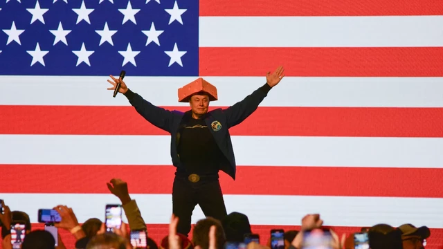 Elon musk standing in from of an american flag with his arms wide and an odd triangle hat that resembles a block of cheese

A small crowd before him with phones out