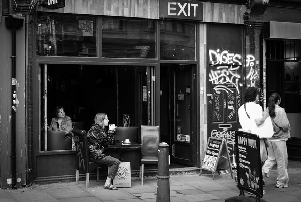 Black and white. The outside of the Exit bar and cafe in London's Brick Lane, with a large window open to the street, an open door on the right, and a sign saying, "EXIT" above the door. A woman is sitting inside looking out and another woman is sitting at a table outside. On the pavement to the right are two signs, one an A-frame saying, "Eat Korean Now Open", the other a vertical sprung sign saying, "Happy Hour", with the times and prices of cocktails, wines, and beer. Further to the right, two women are about to walk out of the scene.