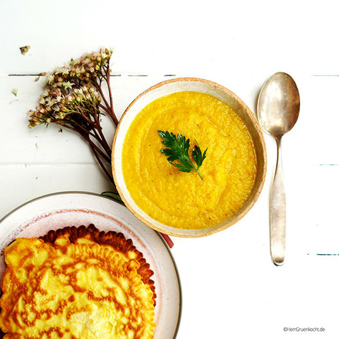 Weißer Holzuntergrund. Darauf eine Suppenschale mit einer Kartoffel-Möhren-Petersilienwurzel-Suppe. Links unten ein Teller mit Pfannkuchen.