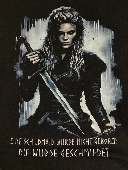 auf dem Bild ist eine nordische Frau, dunkel gekleidet, mit Schwert und Dolch zu sehen, darunter der Spruch:
"Eine Schildmaid wurde nicht geboren"
"Die wurde geschmiedet"