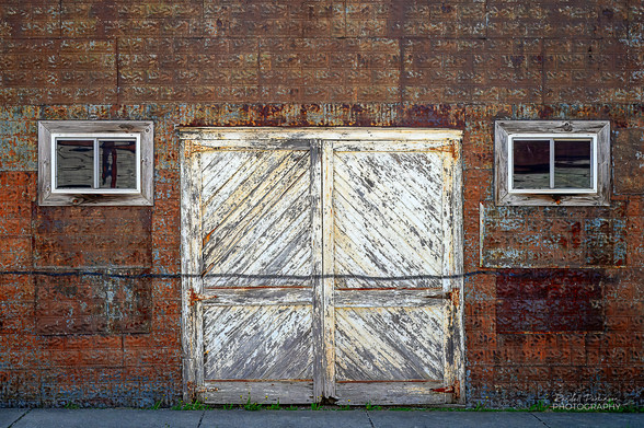 A white, weathered, wood double door and two windows in a building covered with rusty stamped metal siding.