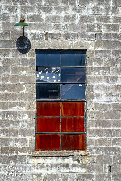 A 12 pane window has 6 lower panes with rusty metal plates replacing glass.  The top six panes have a blue tint from reflecting the sky.  The wall is made of concrete block that at one time may have been painted white.  A single outdoor light is attached just above and to the left of the window.