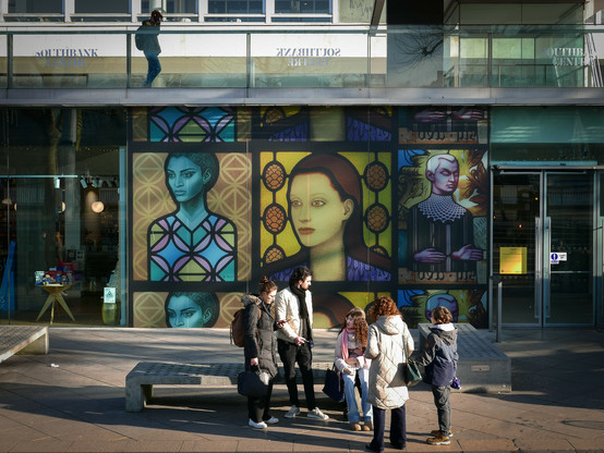 Colour. A family of tourists, one man, three women, a boy and a girl, are sitting on or standing by a concrete bench outside The Royal Festival Hall on London's South Bank. We are seeing them from a few feet above, at the top of some steps. Behind them, on the large windows between two glass-dooredentrances, are a set of nine decals of what looks like illustrations from books, two female faces on the left and middle, and a male face on the right.