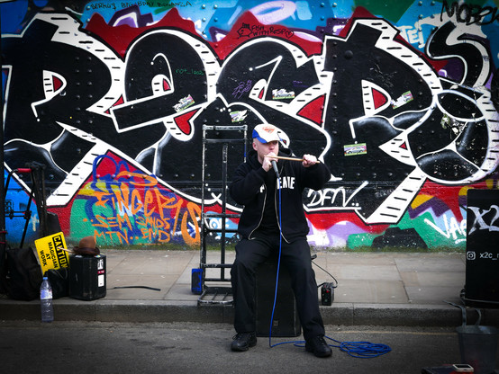 Colour. A beat boxer dressed all in black apart from a blue and white baseball cap holds his mic on his mouth with his right hand and a drumstick in his left hand like a wand. He is sitting on a black case that looks like it holds some of his equipment. On the right of the picture is what looks like a loudspeaker with 'X2' painted on it with part of an Instagram address below it. At the left of the picture is some of his gear including a black box about 14" high on top of which sits a brown fedora hat and another drumstick. To the left of this is a yellow and black sign saying, "Caution, magician at work". In front of this is an 500ml water bottle, possibly used to clean half a gallon of spittle off the mic after the performance. The backdrop is a graffiti-covered wall with the name 'RESP' in black on a colourful background.