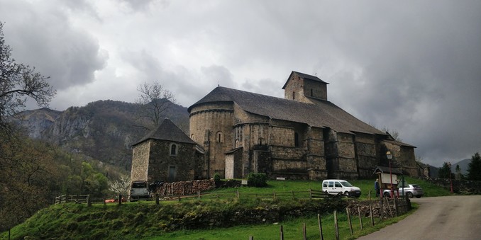 Iglesia románica del siglo XI. Tiene contrafuertes enormes y una nave bastante alta. Es la iglesia románica más grande que he visto nunca, ahí, que no hay más que cuatro casas. En el fondo de ven montes muy altos, la hierba es muy verde y hay nubes muy oscuras y amenazantes.