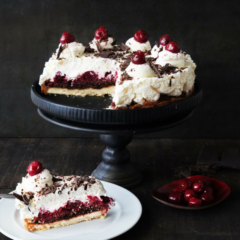 Dunkler Hintergrund. Auf einem Kuchenständer: Eine angeschnittene Schwarzwälder-Kirsch-Tarte. Unten ein Stück davon auf einem Teller.
