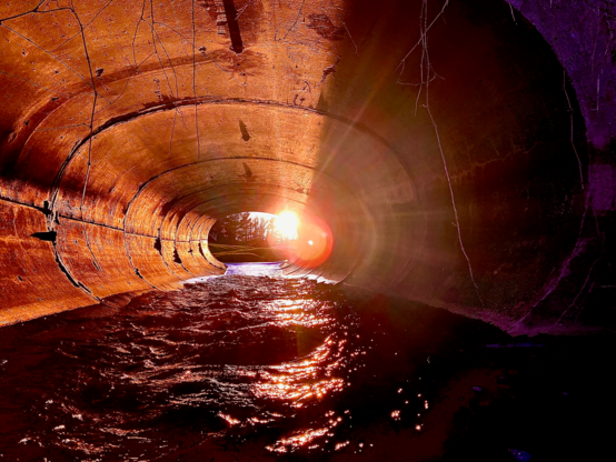 Color image of the moment the setting sun aligns with the culvert/sewer pipe conducting Yellow Creek under a road in a suburban neighborhood, Bath Township, Summit County, Ohio, USA. Image shows the sun directing its beams through the culvert from end to end, lighting up the creek surface and setting the walls of the culvert aglow in shades of orange, yellow, and red.