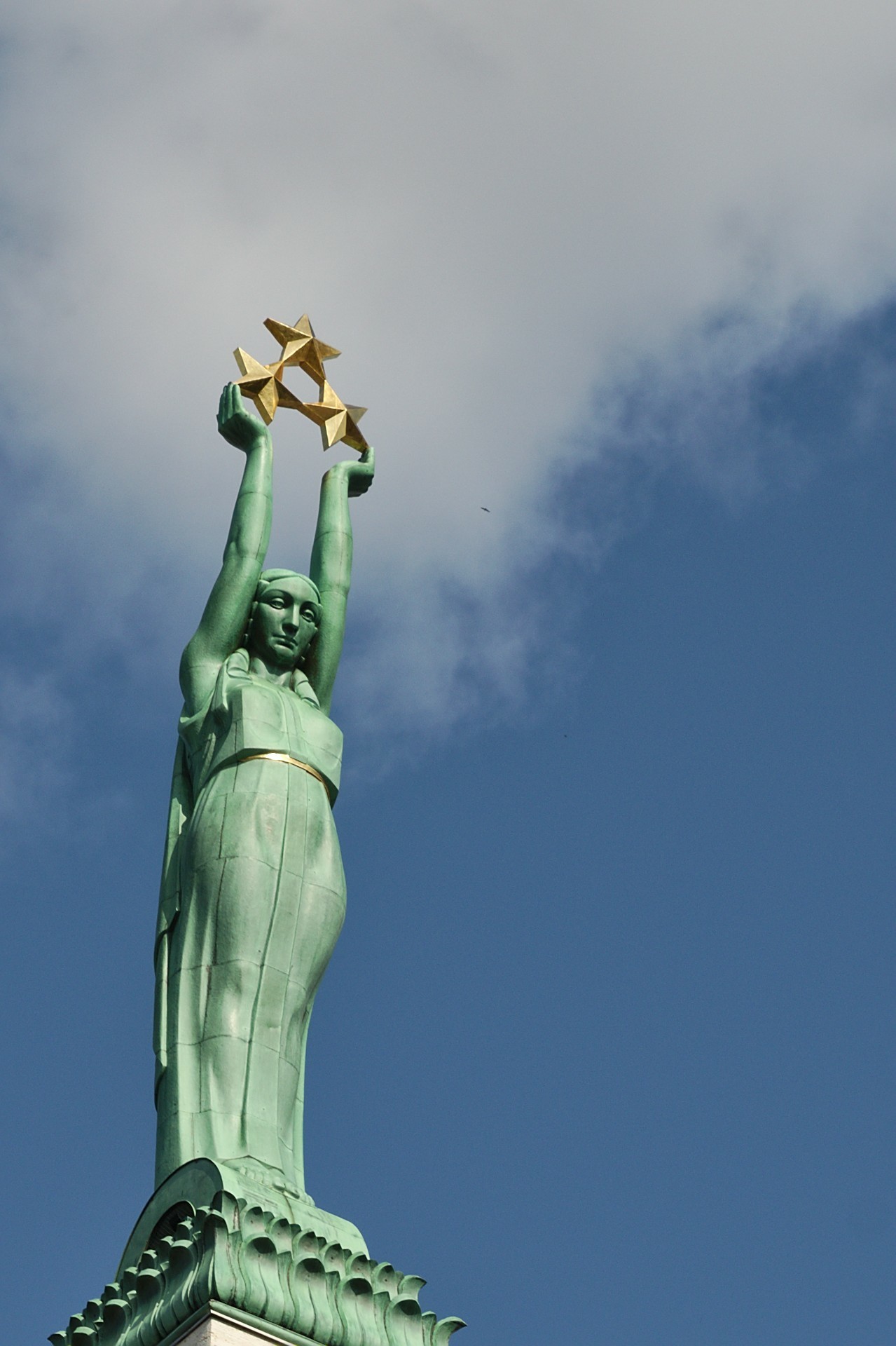 A green copper statue of a woman, arms stretched out straight upwards, holding three goldeen five-point stars in a triangular arrangement. The shot is set against a blue sky with white clouds. A green copper statue of a woman, arms stretched out straight upwards, holding three goldeen five-point stars in a triangular arrangement. The shot is set against a blue sky with white clouds.