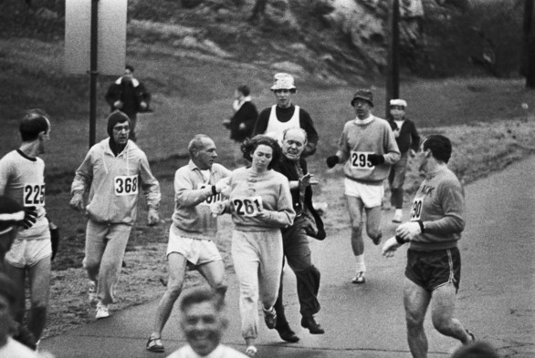 Kathrine Switzer running along a road in a grey tracksuit. She is a white woman with dark hair. There are several other men running alongside her, also in running gear. They are protecting her from a man in a blazer who is attempting to rip off the number pinned to her back.
