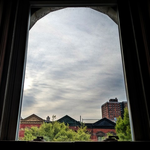 Looking through an arched window ninety minutes after sunrise the sky is full of amorphous , streaky, grey clouds, and the sun is almost breaking through on the left. Pointed roofs of Harlem brownstones with red brickwork are across the street, and a taller apartment building can be seen in the distance. The green tops of two trees are on the bottom and right.