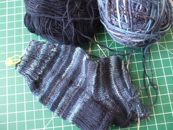 striped socks in progress on a green cutting board. Solid navy yarn alternating stripes with sparkly variegated blue yarn. Heel is solid navy. 