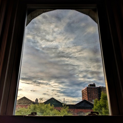 Looking through an arched window an hour after sunrise the sky is full of broken grey and white clouds, the sun is just out of frame on the left, and a patch of blue sky is visible. Pointed roofs of Harlem brownstones with red brickwork are across the street, and a taller apartment building can be seen in the distance. The green tops of two trees are on the bottom and right.