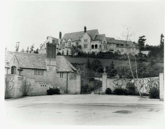 A vintage black and white view of a Tudor-style house high on a hill above the street. 