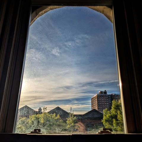 Looking through an arched window an hour after sunrise the blue sky is beatific, with a lovely mix of clouds and warm sunlight from the left side. Pointed roofs of Harlem brownstones are silhouetted across the street, and a taller apartment building can be seen in the distance. The green tops of two trees are entering the frame on the bottom and right. The window is grimy on the bottom.