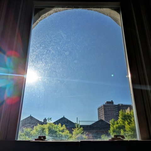 Looking through an arched window 100 minutes after sunrise the sun is above the buildings, making a dramatic lens flare that bisects the frame diagonally with a curved white line and multi-colored petals on the left. The sky is deep blue and cloudless. Pointed roofs of Harlem brownstones are silhouetted across the street, and a taller apartment building can be seen in the distance. The window is grimy. The green tops of two trees are on the bottom and right.