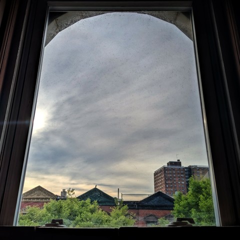 Looking through an arched window two hours after sunrise the sky is full of thin grey-blue clouds, with a patch of light orange sky at the horizon as the sun almost breaks through above the buildings on the left . Pointed roofs of Harlem brownstones with red brickwork are across the street, and a taller apartment building can be seen in the distance. The green tops of two trees are on the bottom and right.