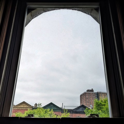 Looking through an arched window two and a half hours after sunrise the sky is full of grey clouds. Pointed roofs of Harlem brownstones with red brickwork are across the street, and a taller apartment building can be seen in the distance. The green tops of two trees are on the bottom and right.
