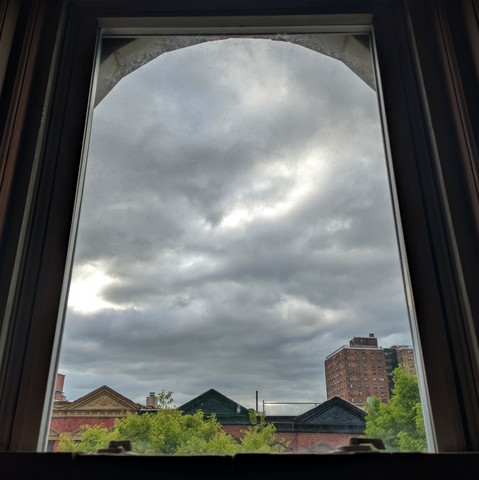 Looking through an arched window two and a half hours after sunrise the sky is full of chunky grey clouds. Pointed roofs of Harlem brownstones with red brickwork are across the street, and a taller apartment building can be seen in the distance. The green tops of two trees are entering the frame on the bottom and right.