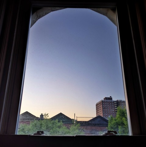 Looking through an arched window at the moment of sunrise the cloudless sky fades from light peach at the horizon to dusty blue up above. Pointed roofs of Harlem brownstones  are silhouetted across the street, and a taller apartment building can be seen in the distance. The green tops of two trees are entering the frame on the bottom and right.