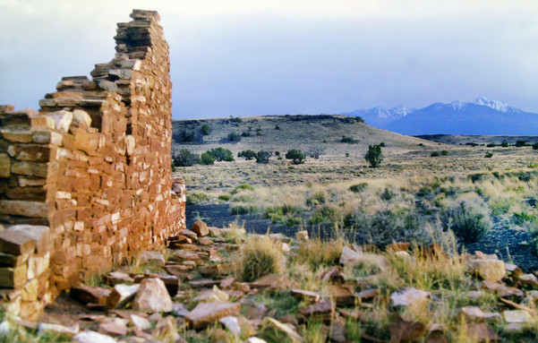 In the golden light of early evening beneath a pale gray blue sky, we are standing in front of partially excavated ancient native American pueblo ruins built of adobe bricks. The semiarid plain beyond is covered with pale gold grasses with occasional green leafy shrubs and a few trees. Numerous rocks and small boulders are visible where the grass does not grow. Small grassy ridges and hills rise in the background. Above and beyond them a range of loftier snowcapped mountains towers in the distance.